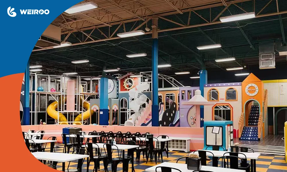 Overview of a modern Family Entertainment Center (FEC) layout, showing a large soft play structure in the background and a cafe seating area for parents in the foreground, illustrating the recommended hybrid business model.