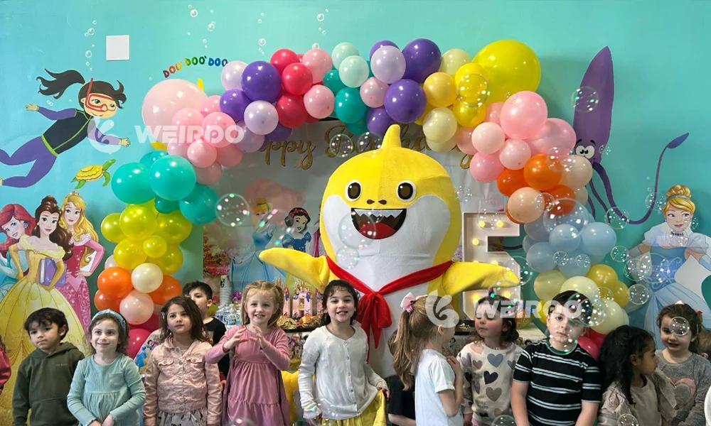 Children celebrating a birthday party at an indoor playground, complete with balloon arches and a shark mascot. This illustrates the high-margin party revenue stream essential to a successful business plan.